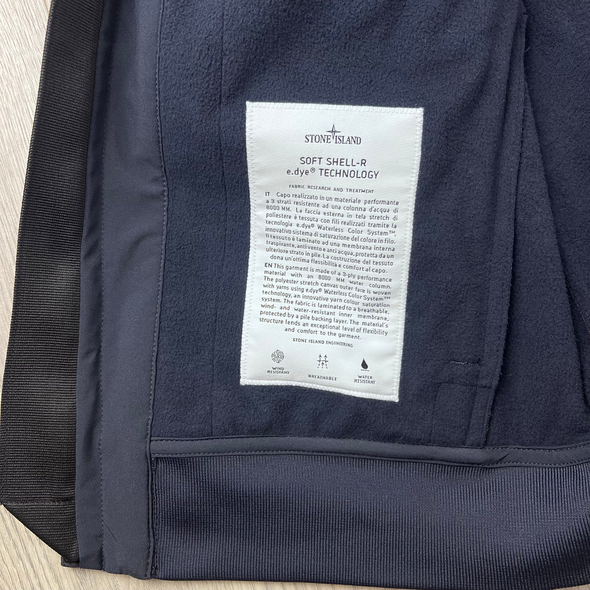 A close up view of the engineering label on the inside of a Stone Island Soft Shell-R_e.dye® Jacket in Navy Blue.